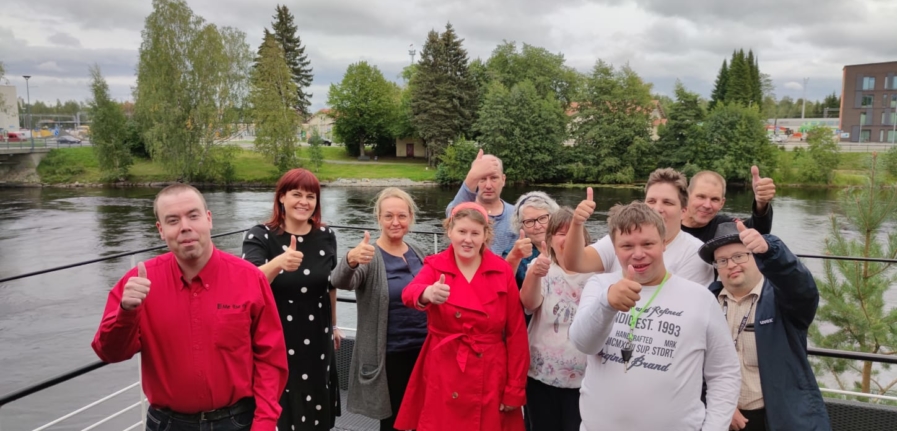 Ryhmäkuva, jossa 11 ihmistä seisoo terassilla toisen käden peukalo pystyssä.