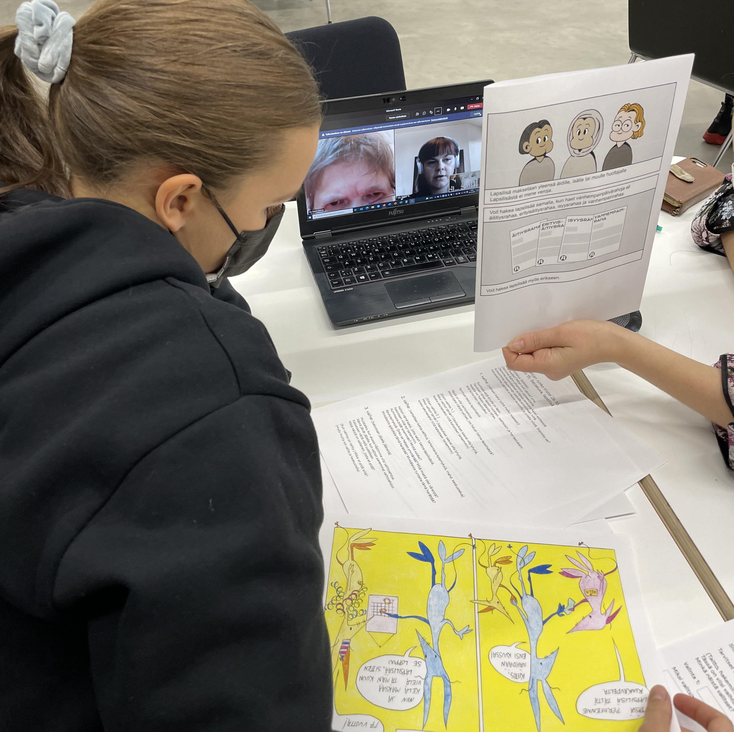 Yksi opiskelija näyttää Teams-kokouksessa olevalle henkilölle sarjakuvaa, toinen opiskelija seuraa sivusta. Kuvassa näkyy myös sarjakuva, jossa hahmot ovat eläimiä.
