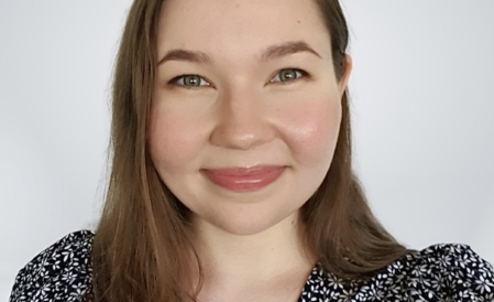 Nuori ruskeahiuksinen hymyilevä nainen vaalealla taustalla.