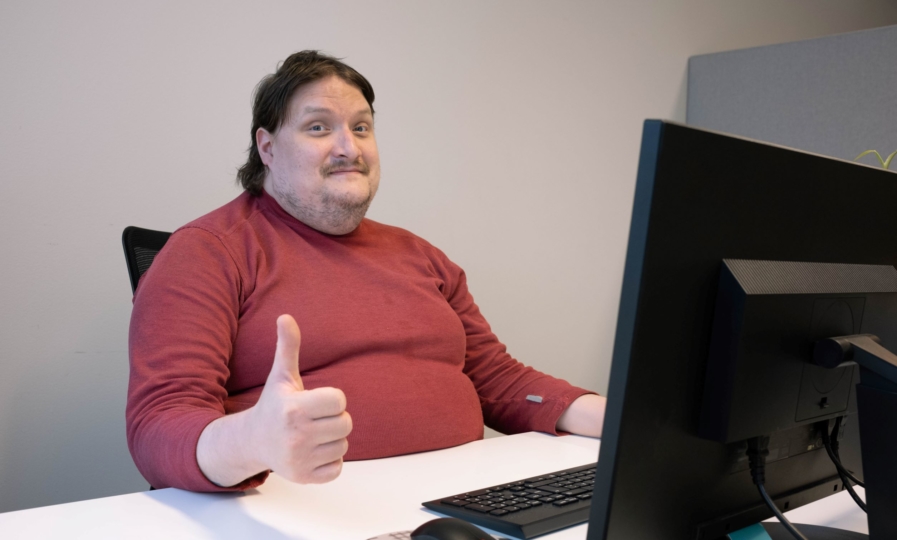 Mies istuu pöydän ääressä tietokoneen edessä ja näyttää kuvaajalle peukkua.